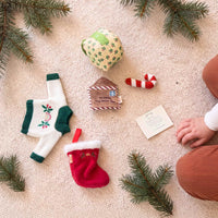 Dinkum Doll Christmas accessories laid on a cream knit rug with pine sprigs: a knitted holly jumper, red stocking, plush gift box, candy cane, letter to Santa and a note card.