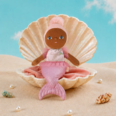 A mermaid doll with brown skin, blue eyes, and pink hair sits inside a large beige clam shell on sand, with a turquoise sky backdrop and small shells around it.