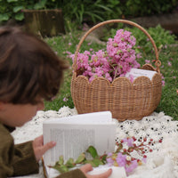 Natural rattan Petal Basket with a scalloped edge and sturdy handle filled with pink flowers beside a child reading outdoors on a blanket