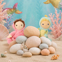 Two plush mermaid dolls sitting on a pile of smooth beach stones, with coral, shells, and a blue underwater-themed backdrop.