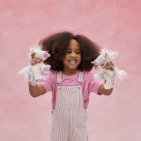 Smiling child holding two soft white unicorn plush toys with pastel details against a pink background from Dinkum Pets Unicorn Collection