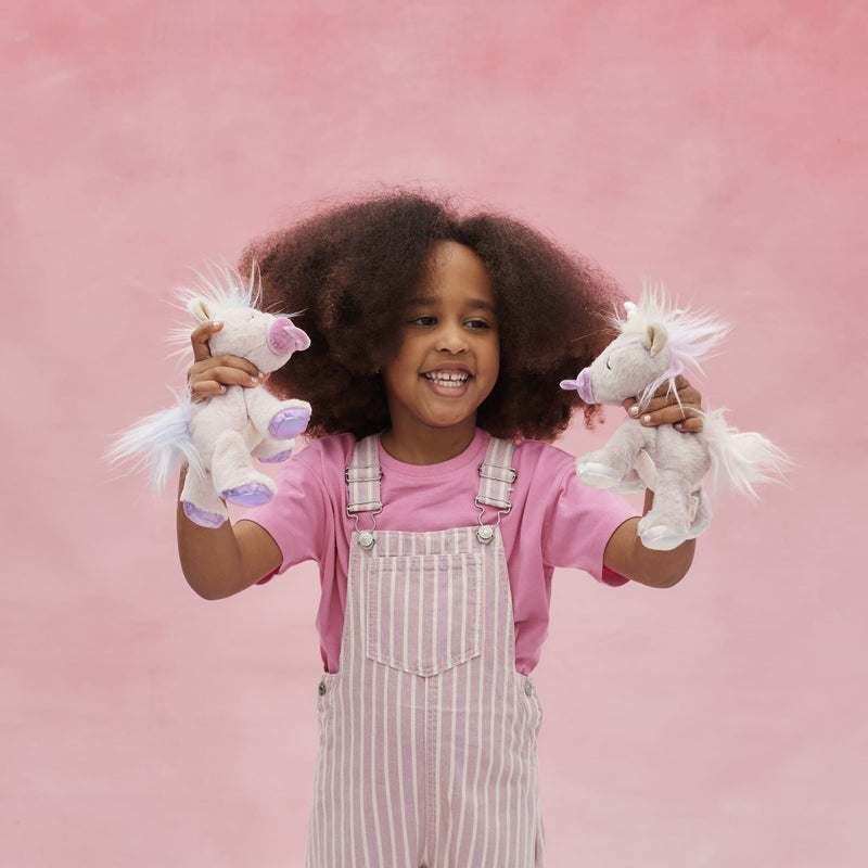 Smiling child holding two soft white unicorn plush toys with pastel details against a pink background from Dinkum Pets Unicorn Collection