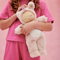 Child in pink clothing cuddling a soft blush Cozy Dinkums unicorn plush with a sleepy embroidered face fuzzy ears and a twinkling purple horn