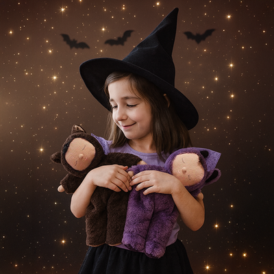 Child wearing a witch hat cuddles two soft Cozy Dinkum dolls including Bat Pip against a sparkling night sky with flying bats.