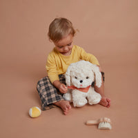 Child cuddles a fluffy white Dinkum Dogs plush puppy with a coral bandana while a yellow ball and grooming brush sit nearby for imaginative play