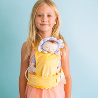 Child wearing a yellow Dinkum Dolls Petal Carrier with a sleeping doll in a purple bonnet against a soft blue background
