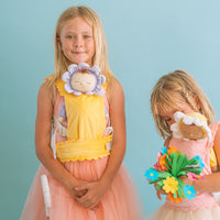 Child wearing a yellow Dinkum Dolls Petal Carrier with a flower faced doll while another child holds a doll and colourful bouquet during imaginative play