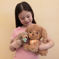 Child cuddling two soft Dinkum Dogs plush toys including a golden dog with an orange bandana and a smaller brown dog with a mint green collar