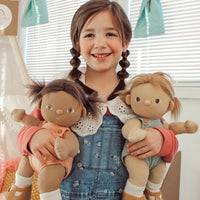 A smiling child holds two soft Dinkum Dolls with brown hair and cosy outfits in a bright room for imaginative play