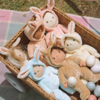 Six Dinky Dinkums bunny dolls in pink blue cream and brown outfits nestled in a wicker basket with pastel Easter eggs on a picnic blanket.