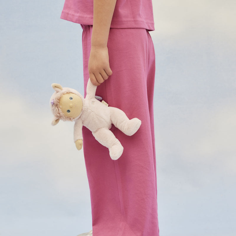 A child holds a soft cream Dinky Dinkums unicorn doll with a pink mane and embroidered face against a pale blue background