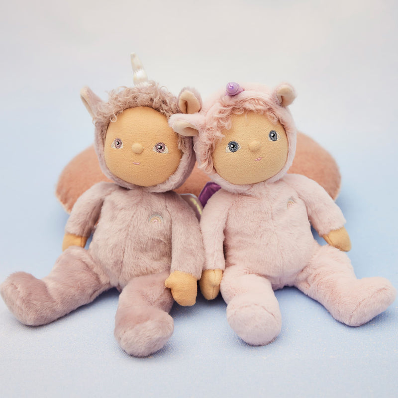 Two soft pink Dinky Dinkums dolls sitting together with cuddly plush bodies and sweet embroidered faces one featuring a unicorn horn and the other a flower hair detail