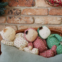 Three sleeping Dozy Dinkum dolls nestled together in a woven basket under a soft blanket against a rustic brick wall by Dinkum Dolls by Olli Ella