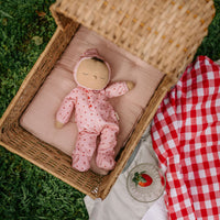 Dozy Dinkums Pip Strawberry soft doll in a pink strawberry print sleeps in a wicker basket beside a red checked picnic blanket