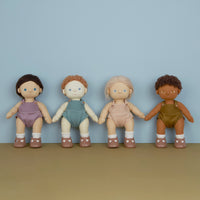 Four Dinkum Dolls with diverse skin tones and hairstyles stand hand in hand wearing pastel rompers white socks and brown shoes against a blue backdrop