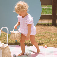 Child in a pink outfit carries a small handwoven rattan basket with a floral liner during outdoor play on a picnic blanket.