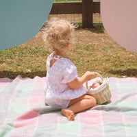 Child sitting on a pastel blanket outdoors holding a small woven rattan basket with a cream floral liner from Olli Ella Kids Rattan