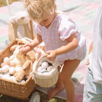 Child holding a small Olli Ella rattan basket with a cream floral liner beside a toy pram filled with soft bunny toys and pastel eggs outdoors