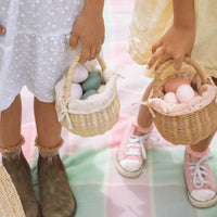 Two children carry handwoven rattan baskets lined with floral cotton fabric and filled with pastel play eggs from Olli Ella Kids Rattan