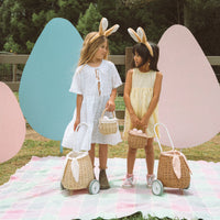 Two children wearing bunny ears play outdoors with small rattan baskets and wheeled doll prams on a pastel picnic blanket by colorful garden panels