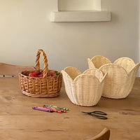 Buttercream rattan basket set by Olli Ella Nursery displayed on a wooden table with woven handles and a second basket filled with flowers nearby