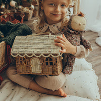 A smiling child holds a rattan gingerbread house clutch with a woven roof and arched door beside a soft doll in a festive playroom