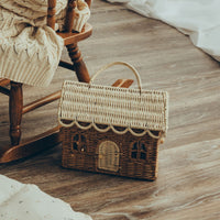 Olli Ella rattan gingerbread house clutch with woven handle resting on a wooden floor beside a rocking chair and soft knit blanket