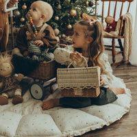 Child holding a rattan gingerbread house clutch beside a doll and baby during festive play near a decorated Christmas tree.