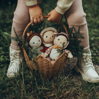 Child carrying a woven natural rattan basket filled with three soft dolls and festive greenery outdoors. The basket has a scalloped edge and sturdy handle.
