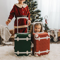 Two children beside forest green and red rolling suitcases in a cosy Christmas setting with a decorated tree and festive gifts by the window