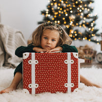 Child resting behind a Sweetheart Red polka dot suitcase by Olli Ella Kids beside a decorated Christmas tree at home during the holiday season