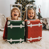 Two smiling children pose behind colorful Olli Ella Kids See Ya Suitcases beside a decorated Christmas tree with the red polka dot suitcase in front