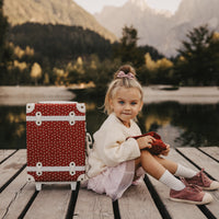 Olli Ella Kids red polka dot suitcase beside a young girl sitting on a wooden dock by a mountain lake with a small red bag