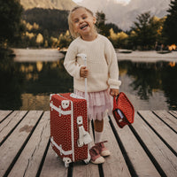 Smiling child holding a red patterned wash bag beside a matching suitcase on a lakeside dock with mountains in the background by Olli Ella Kids