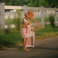 Child walking outdoors with a pink strawberry print kids suitcase and a doll in a sunny garden setting from Olli Ella Kids