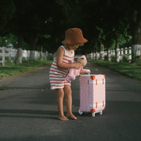 Child in a striped dress and sun hat playing with a doll beside a pink strawberry print suitcase on a tree lined path from Olli Ella Kids