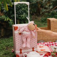 Pink strawberry print children's suitcase with an extended handle and a doll in matching pink clothing at an outdoor picnic by Olli Ella Kids