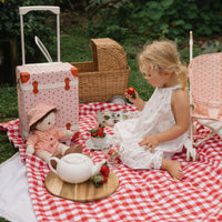 Child enjoys a picnic with a doll and Olli Ella Kids strawberry suitcase on a red gingham blanket outdoors