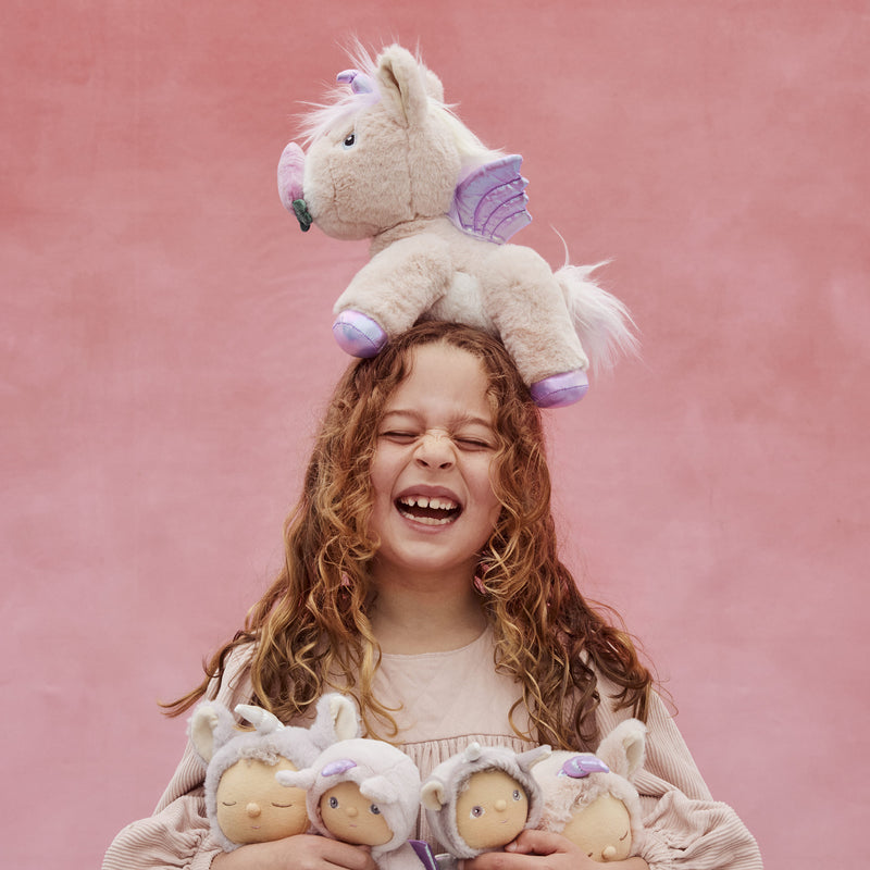 Laughing child holding several soft dolls with a plush unicorn balanced on her head against a pink backdrop from Dinkum Pets imaginative play collection