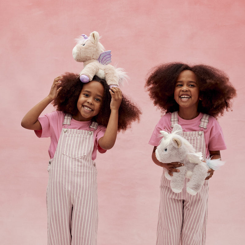 Two smiling children in pink striped overalls play with soft white plush unicorns against a pink backdrop from Dinkum Pets