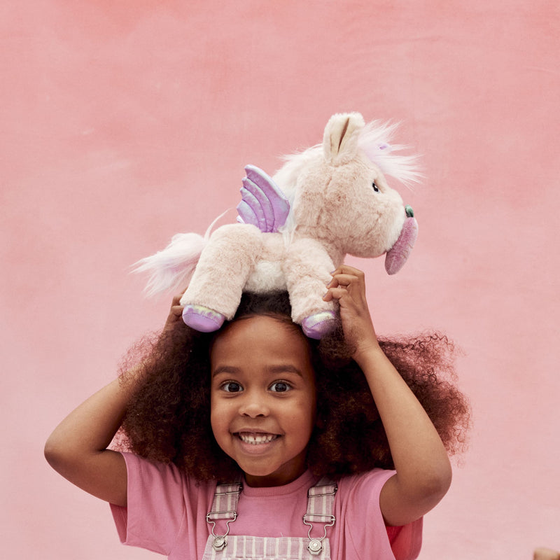Child smiling while holding a soft cream plush unicorn with pink details and purple wings above her head against a pink background from Dinkum Pets