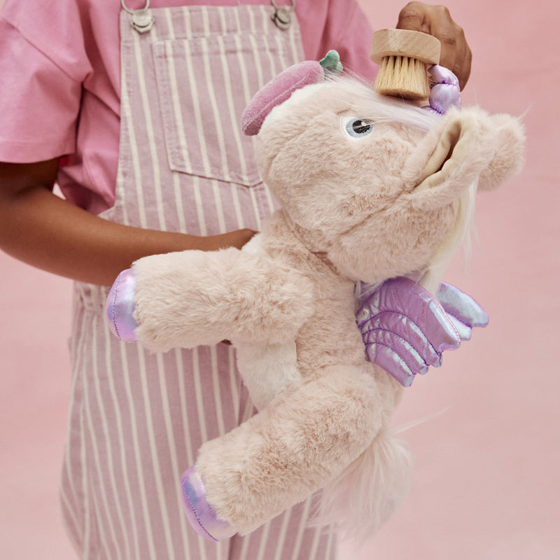 Dinkum Pets plush pink unicorn toy with lilac wings and hooves being brushed by a child in a striped apron during imaginative play