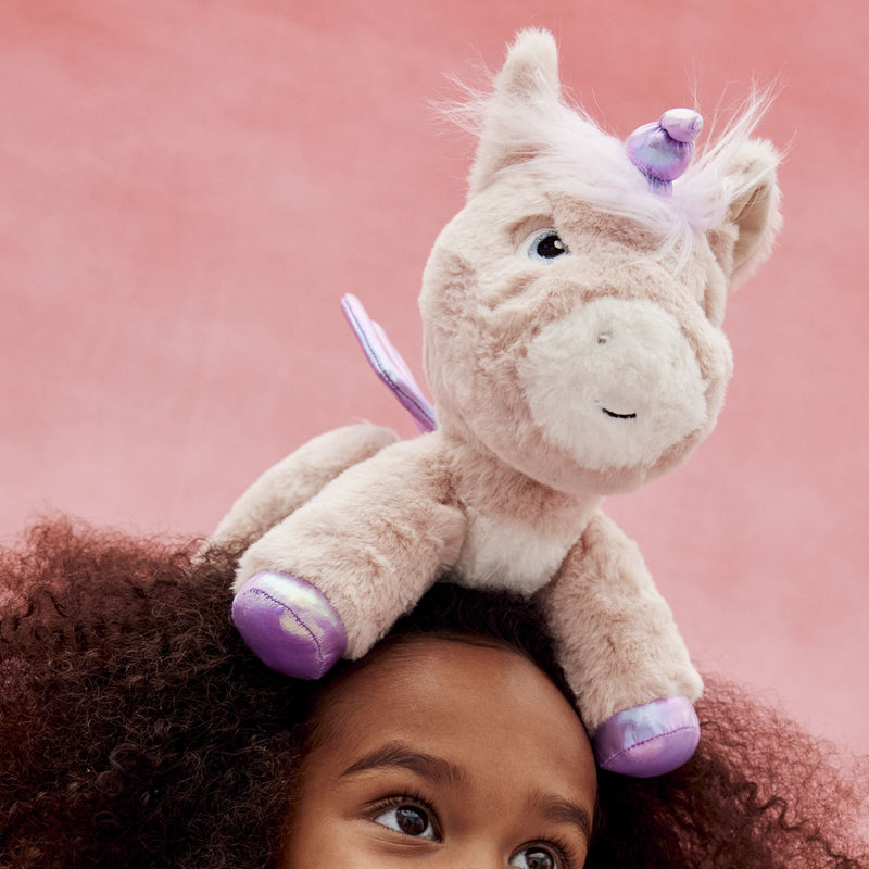 Plush pink and cream unicorn toy with purple details perched playfully on a childs head against a soft pink background from Dinkum Pets
