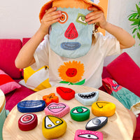 Child plays with Olli Ella Tubbles Funspressions sensory stones featuring expressive facial shapes on a table while holding a matching blue pouch