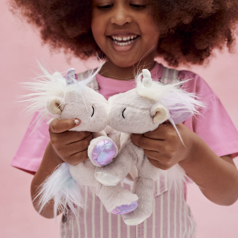 Smiling child holding two soft white and lilac plush unicorns with glittery horns and fluffy manes from Dinkum Pets against a pink background