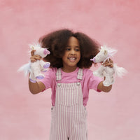Smiling child in pink overalls holding two soft white unicorn toys with lilac details and flowing manes against a pink backdrop
