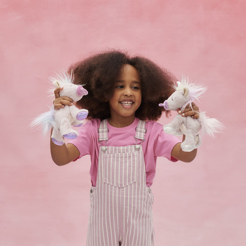 Smiling child in pink overalls holding two soft white unicorn toys with lilac details and flowing manes against a pink backdrop