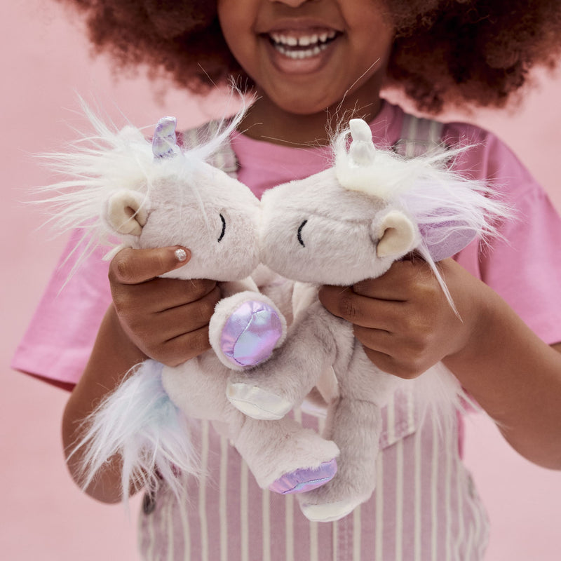 A smiling child holds two soft white unicorn plush toys with pastel pink and lilac details against a pink background from Dinkum Pets