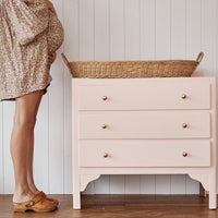 Olli Ella Nursery Bayu rattan changing basket with leather handles resting on a blush pink nursery dresser beside a parent in a floral dress