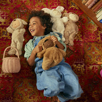 A smiling child cuddles a soft brown doll beside three plush dolls and a rose rattan basket on a patterned rug from Olli Ella Home